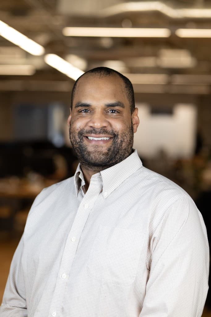 Professional headshot of Kevin Fich smiling, wearing a light-colored checkered shirt in a blurred office setting.